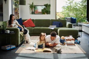 A man reading with a young girl with a woman perched on a couch watches on. 